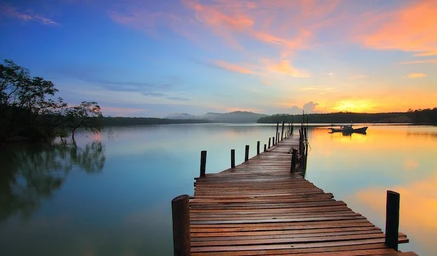 Photo of sunrise at end of a dock.Blue water, orange and pinks in sunrise.  Photo by Sabri Ismail in Malaysia via Pixabay