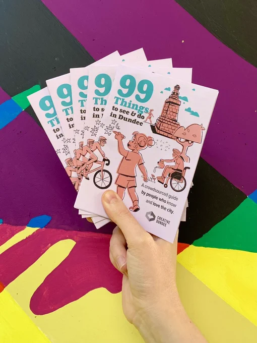 Colourful painted background. A hand is seen holding a stack of printed 99 Things to See & Do in Dundee Guides.