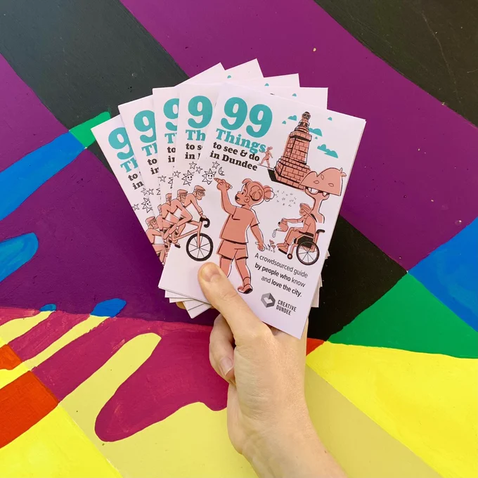 Colourful painted background. A hand holds a stack of printed Dundee Guides.