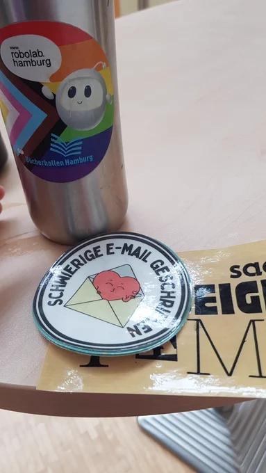 Im Vordergrund: Badge, auf dem steht: #Schwierige E-Mail geschrieben" über einem beigen "Sad beige Fami" - Sticker. Im Hintergrund Thermosbecher mit Kaffee