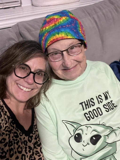 Two white women with glasses in pajamas sitting on a gray couch. One is wearing a rainbow tie dye head scarf and a pale green Grogu shirt. The other has chin length dark hair.