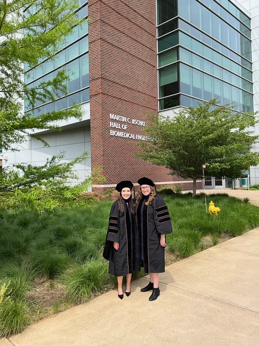Two women stand in front of the Martin C Jischke Hall of Biomedical Engineering. They are wearing graduation regalia and smiling.