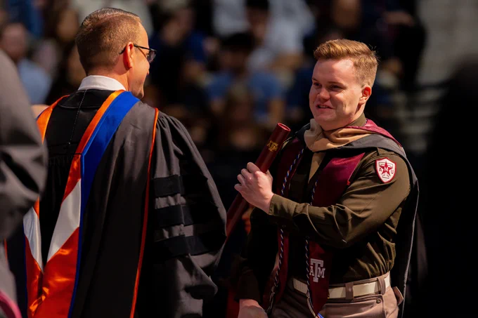 A graduate wearing a Corps uniform gripping his diploma and smiling on stage.