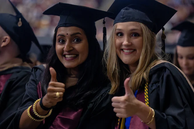 Two graduates seated in the crowd gigging 'em and smiling next to each other.