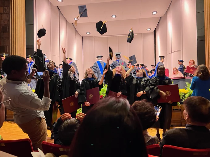 six doctoral students in their regalia throwing their caps into the air to celebrate graduation after the ceremony