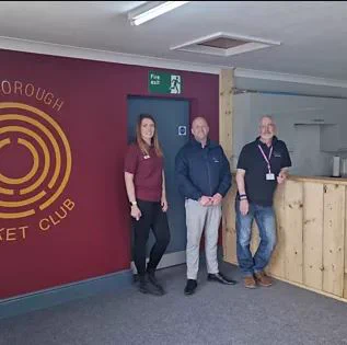 Three people stood in a reception area in front of a wall with a logo for Alkborough cricket club.
