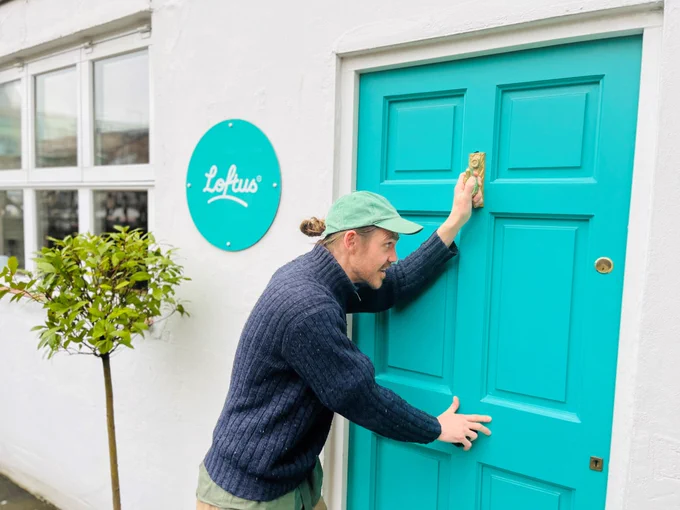 actor Jack Parris knocks and leans on a turquoise coloured front door with the company name 'Loftus' on a sign on the left. 