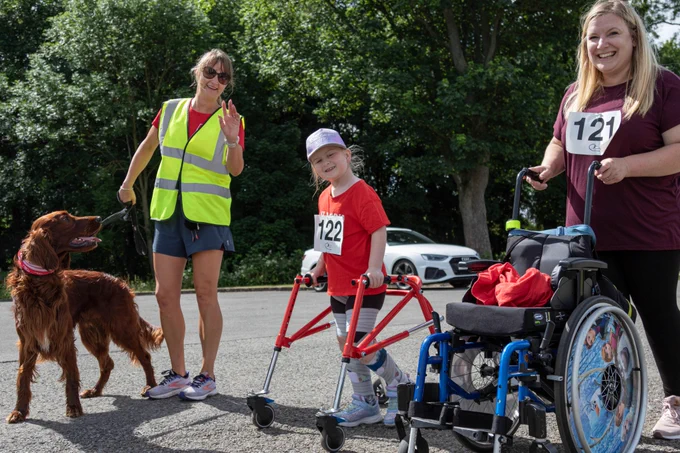 A young girl completing Run with Rich in her walking frame alongside a woman pushing a wheelchair and a fantastic volunteer and her dog. All three people are smiling and looking at the camera