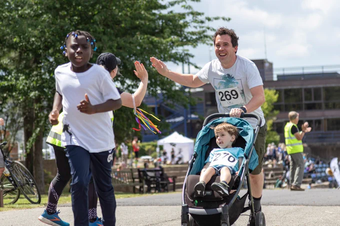 A young child in the foreground is running towards the camera smiling; behind him is a man pushing a running buggy with a young boy, the man is smiling and high fiving a volunteer.