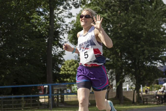 a woman is running with a smile on her face and waving towards the camera.