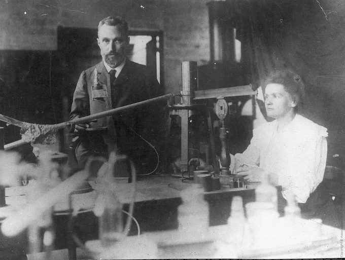A black and white photograph of Pierre Curie (left) and Marie Skłodowska–Curie (right) in their lab.