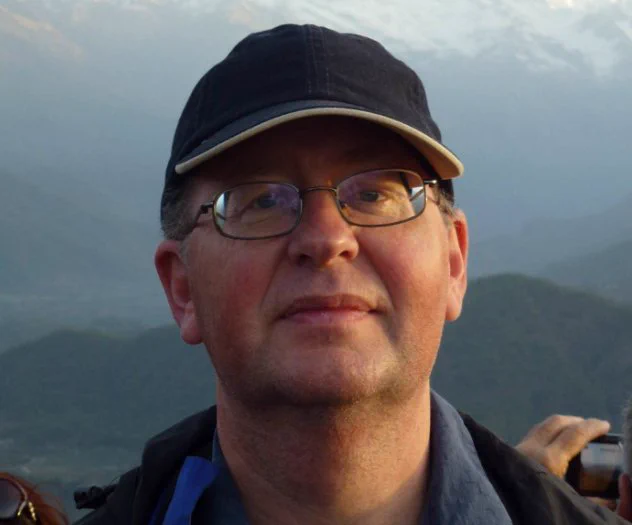 A picture of Jonathan wearing glasses, a dark grey baseball cap with mountains in the background.