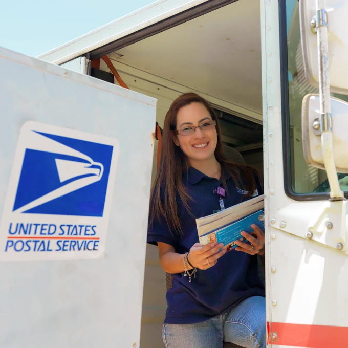 A female letter carrier holds letters while seated in a USPS vehicle next to mailbox with United States Postal Service logo on the side