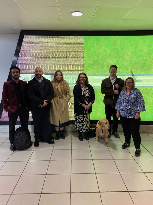 Image description: group photo of the transreport and Heathrow team. There are three males and three females standing in front of a screen with a green background that says 'Heathrow Express'. Tot the left of the photo we have Adam, Joseph and Sophie. In the middle of the photo is Emma, standing next to Luna, her assistance dog. To the right of the photo, we have Jacob and Claire.