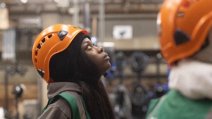 A young woman wears an orange hard hat and a grey hoodie with a high vis green jacket over the top and looks up. In the foreground the edge of another person is visible who wears the same hard hat and high vis jacket. The backdrop is a blurred warehouse setting with cables hanging on the wall.