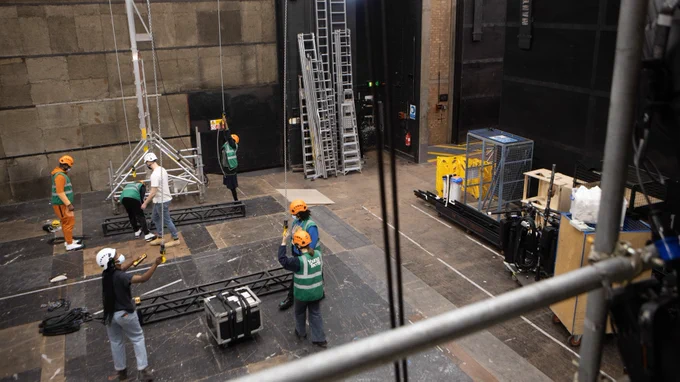 A warehouse type space that is backstage of a theatre, the image is taken high up from a balcony. 5 young people wear orange hardhats and green high visibility jackets. There is a man in a white t-shirt and white hard hat and a woman in a black top, jeans and a white hard hat. There are two lighting bars on the floor amongst them. They hold various cables from the ceiling with and clip them onto the bars. Around them are ladders and huge cardboard boxes etc.