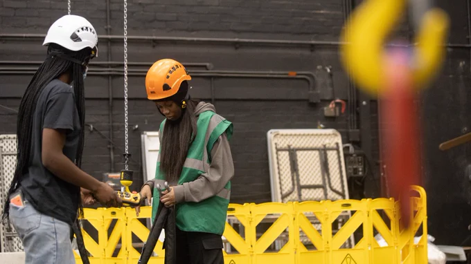 Two young women. One wears a grey hoodie, green high vis jacket and orange hard hat who holds the end of a pipe in her hand. The other, who wears a white hard hat, black t-shirt and jeans, holds a yellow hook that is at the end of a chain hanging from the ceiling. Behind them is a yellow plastic warning fence with fold up tressel tables behind it.