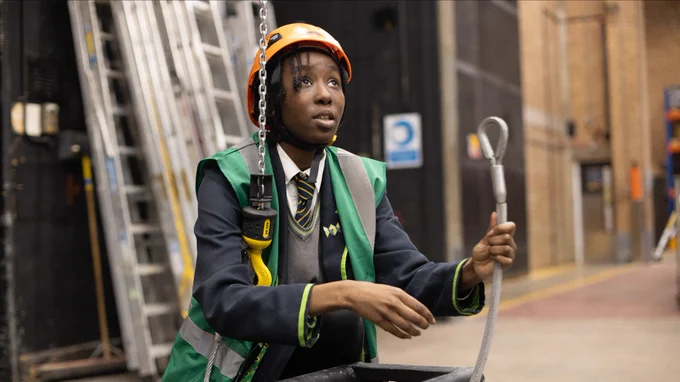 A girl in school uniform, a green high vis jacket and orange hardhat holds a cable with a loop in her left hand. Under her right armpit she tucks a yellow hook that is attached to a chain that comes in from the top of the image as if hung from the ceiling. Behind her there is a stack of roughly 20 ladders. She is in a warehouse type space and looks up at something intensely.