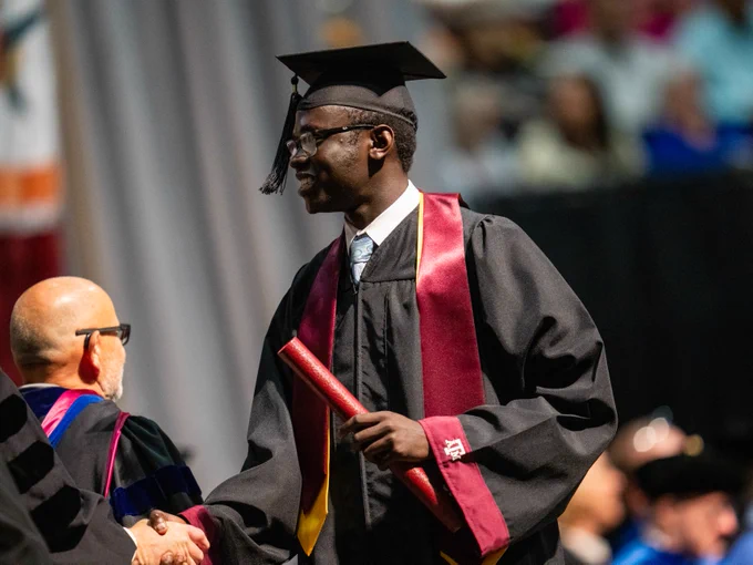 A graduate shaking hands and holding his diploma on stage.