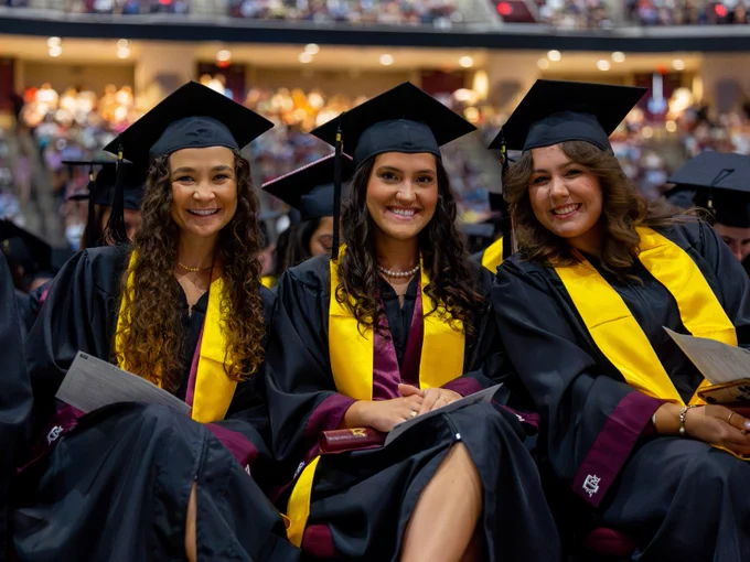 Three graduates seated and smiling for the camera on the floor of the arena.