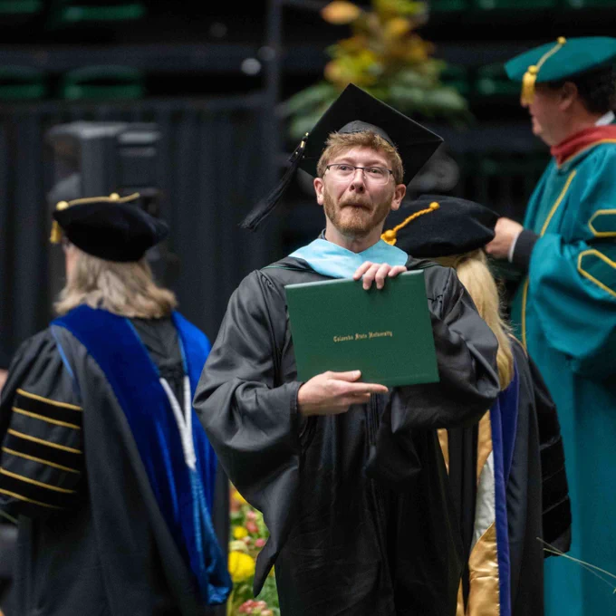 A graduate smiles across the main stage as he holds his degree.
