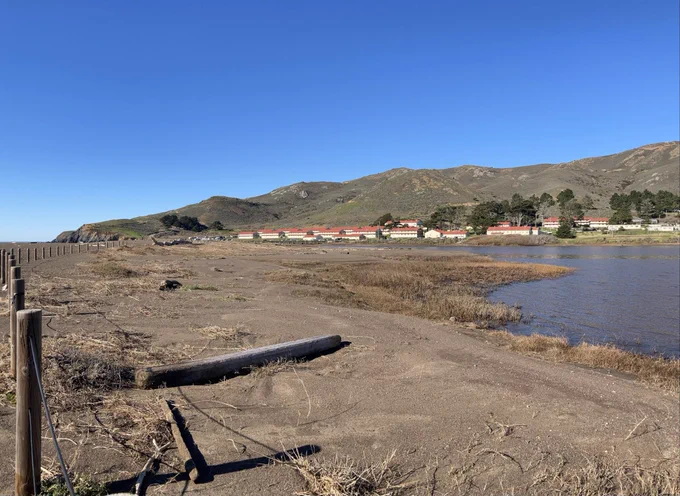 a series of photos of the Rodeo Lagoon taken about 24 hours apart. The before photos show a high-water line up the protective fence in some locations. The after photos show a drop in the water level, and a starkly receded shoreline revealing about 15 feet of dirt and dried grass. In all the photos you can see the rows of old army barracks making up Fort Cronkhite, and rocky, shrub covered hills in the background.