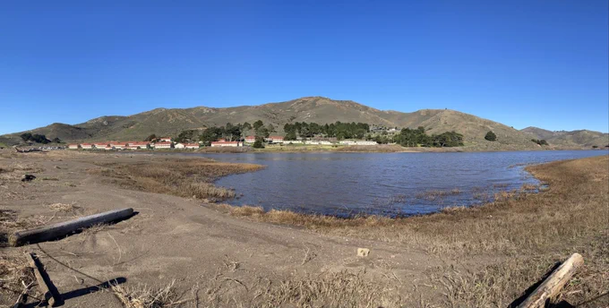 a series of photos of the Rodeo Lagoon taken about 24 hours apart. The before photos show a high-water line up the protective fence in some locations. The after photos show a drop in the water level, and a starkly receded shoreline revealing about 15 feet of dirt and dried grass. In all the photos you can see the rows of old army barracks making up Fort Cronkhite, and rocky, shrub covered hills in the background.