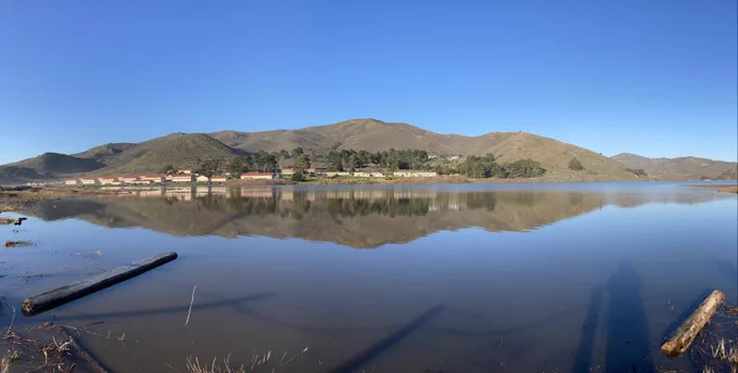 a series of photos of the Rodeo Lagoon taken about 24 hours apart. The before photos show a high-water line up the protective fence in some locations. The after photos show a drop in the water level, and a starkly receded shoreline revealing about 15 feet of dirt and dried grass. In all the photos you can see the rows of old army barracks making up Fort Cronkhite, and rocky, shrub covered hills in the background.