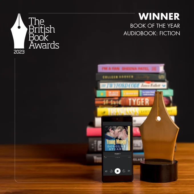 A mobile phone, with the audiobook Young Mungo by Douglas Stuart open on the screen, is upright on a wooden table standing next to a British Book Awards prize in the shape of a pen nib. Against a black background is a pile of books that is slightly blurred. White text at the top reads The British Book Awards, Winner, Book of the Year - Audiobook: Fiction