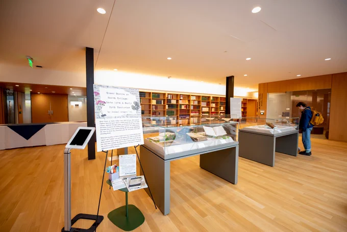 An easel stands next to four glass enclosed display tables containing books, papers, and other pieces collected for student capstone presentations.