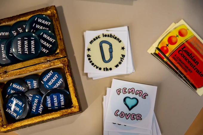 Buttons reading "I WANT", and stickers reading "masc lesbians deserve the world," "femme lover," and "lesbian concentrate: a lesbianthology 100% undiluted" are placed upon a table as part of a student's capstone presentation.