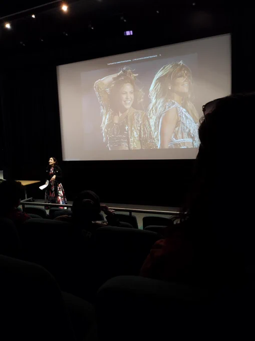 A cinema screen showing J.Lo and Sharika dressed in silver and gold outfits. 
With Mariana standing in front talking to the audience. 