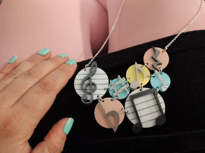 A lap with pale baby pink tights, a hand with mint green nails, a black cord dress and a necklace on top which is a series of circles with musical notes and notations. 
There is a mixture of colours including white, mint green, baby pink, pale yellow and sparkly silver..