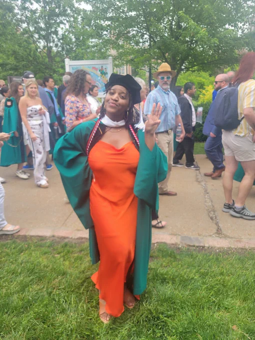 The focus is a black woman wearing an orange dress. She has on a green graduation gown and master's hat. She's holding up 3 fingers to signify completing her 3rd degree