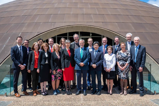 19 men and women stand in front of a dome-shaped building. 