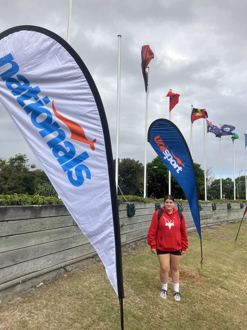Annabelle Stoke standing beside a UniSport Nationals flag wearing her Murdoch Vikings hoodies. 