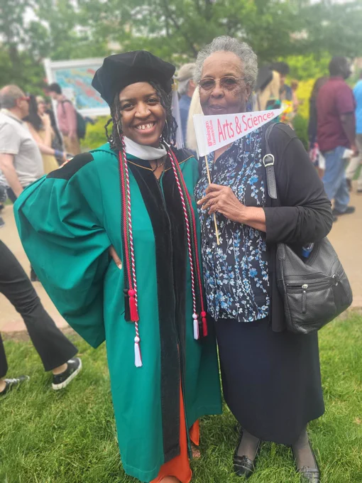 Two black women, one is younger, she's smiling and staring a Tam, graduation gown and hood. The other black woman is older and has short grey hair. She's holding a sign that says Washington University in St. Louis Arts and Sciences