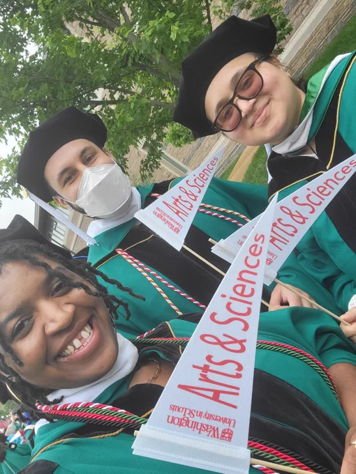 3 graduating students wearing green graduation gowns and black tams. They are all smiling. From left to right, one is a,black woman with locs. The second is a Mexican man wearing a white mask. And the other is an Ecuadorian woman wearing glasses and smiling.