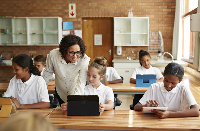 A teacher stands looking at the work of a school student sitting at a desk in a science lab.  The student is working on a tablet, and there are two students next to her.