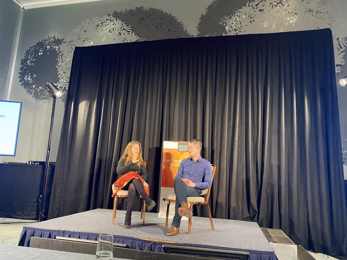 Two people sitting on stage at conference talking with each other. Signage: Get together. Go faster. DevOps Enterprise Summit.