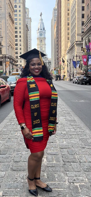 Tonya Bennett standing on a median in the middle of a big street with the city hall building in the background. She is wearing a red dress and  a black, red, gold, green honor stole.