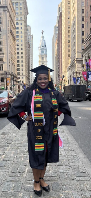 Tonya Bennett standing on a median in the middle of a big street with the city hall building in the background. She is wearing a cap and gown and honor stoles.