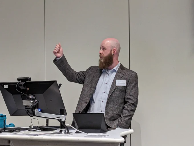 Kris Barr stands behind a PC lectern, with a raised hand while he presents. He has a shaved head, with medium length beard, and wears a grey jacket over a light blue shirt.