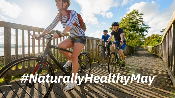 3 people cycling on a wooden bridge. Text reads: “Naturally Healthy May”.
