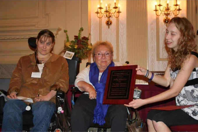 Sally Johnston seated in her wheelchair wearing blue scarf with two other women who are presenting a plaque of the first annual Sally Johnston Advocacy Award.
