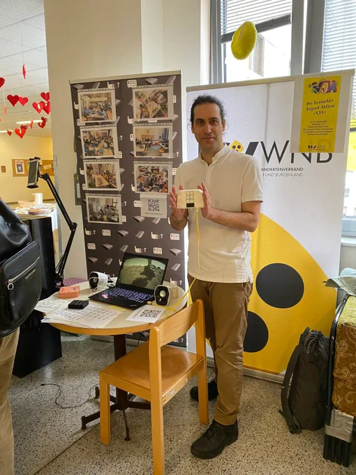 Ein Mensch präsentiert eine mobile Braille-Tastatur und blickt in die Kamera. Seitlich steht ein runder Tisch mit Laptop, Aktivlautsprechern, einer mobilen Braille-Tastatur, ausgedruckten Braille-Alphabeten und einem Smartphone auf einem Schwenkarm befestigt. Unter dem Tisch steht ein Sessel und dahinter zwei Roll-ups. Auf dem linken Roll-up sind Fotos in Klarsichtfolien aufgeklebt und der zweite Roll-up ist vom Blindenverband (BSV-WNB) mit einem zusätzlichen Plakat von der Verrückte-Jugend-Aktion (VJA). Im Hintergrund befinden sich Fenster und von der Decke hängende rote Herzen.