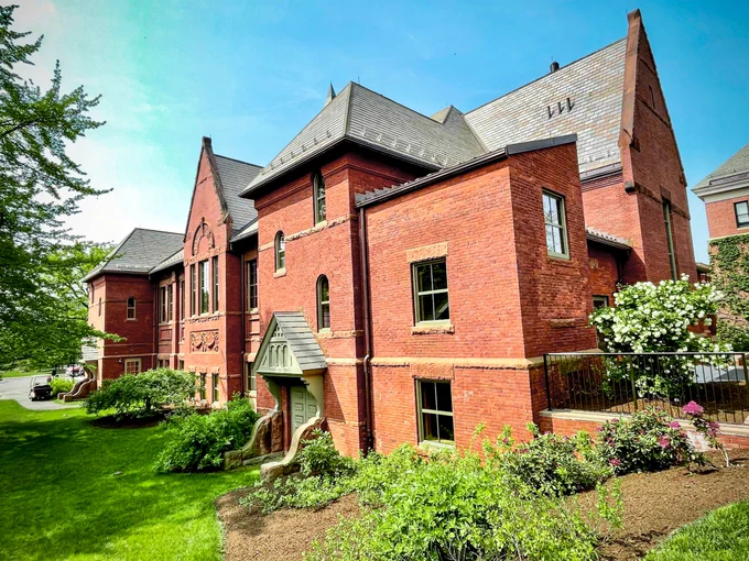 Exterior view of Alumnae Gymnasium on Smith College campus, with blue skies, green grass, and trees and bushes in full bloom.
