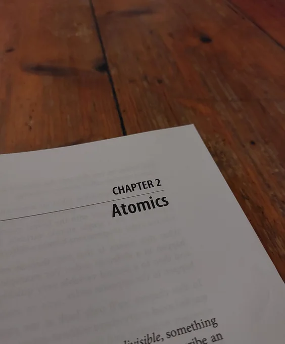 Corner of book page reading 'Chapter 2 - Atomics' on a wooden floor