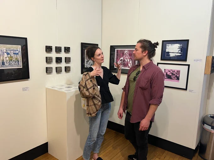 Woman in black shirt and jeans (Grace Carey) speaks with man in unbuttoned burgundy shirt with green t-shirt and black pants in front of framed art. 