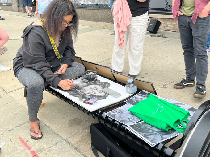 Artist (Monica Redmond) with medium length dark hair wearing hoodie and grey leggings draws a charcoal portrait of a female pop star on a bench. 